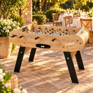 Baby-foot René Pierre Color en bois clair avec pieds noirs inclinés, installé sur une terrasse en pierre dans un jardin de réception élégant.