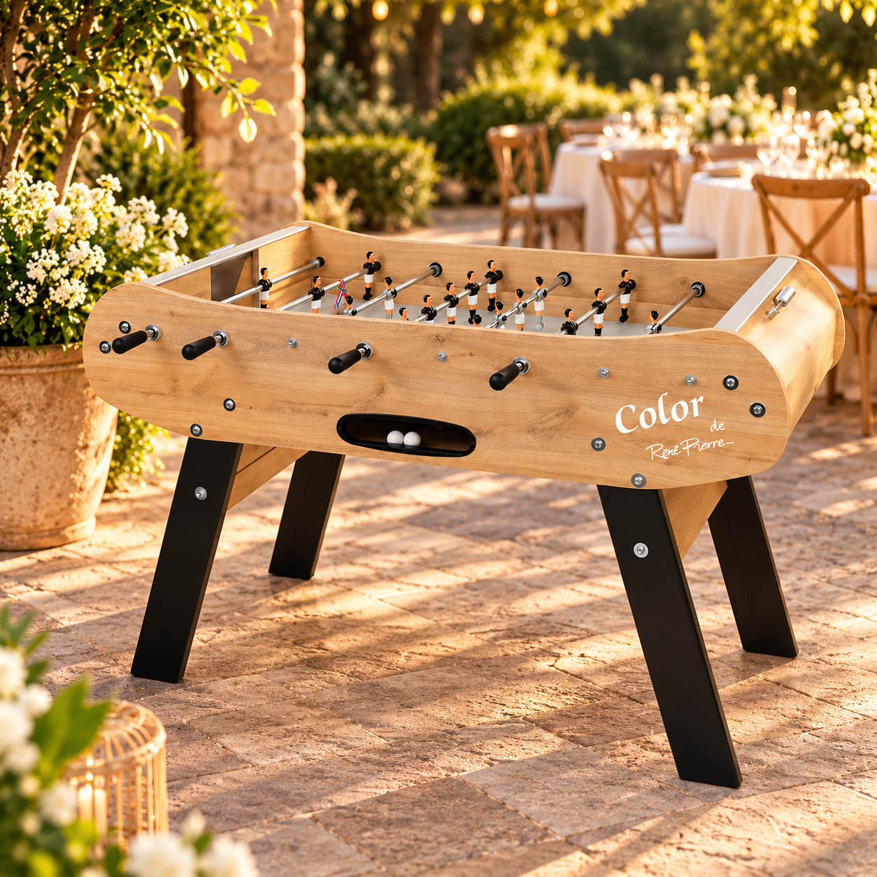 Baby-foot René Pierre Color en bois clair avec pieds noirs inclinés, installé sur une terrasse en pierre dans un jardin de réception élégant.